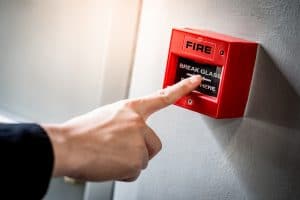 Male hand pointing at red fire alarm switch on concrete wall in office building. Industrial fire warning system equipment for emergency.